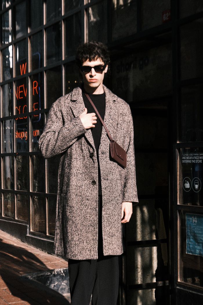delivery-policy Stylish male model in coat, sunglasses, posing in urban area with window reflections.