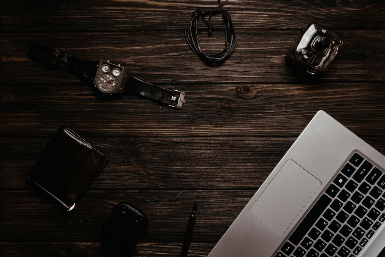 hero-img Flat lay of laptop, watch, wallet, and accessories on a dark wooden surface.